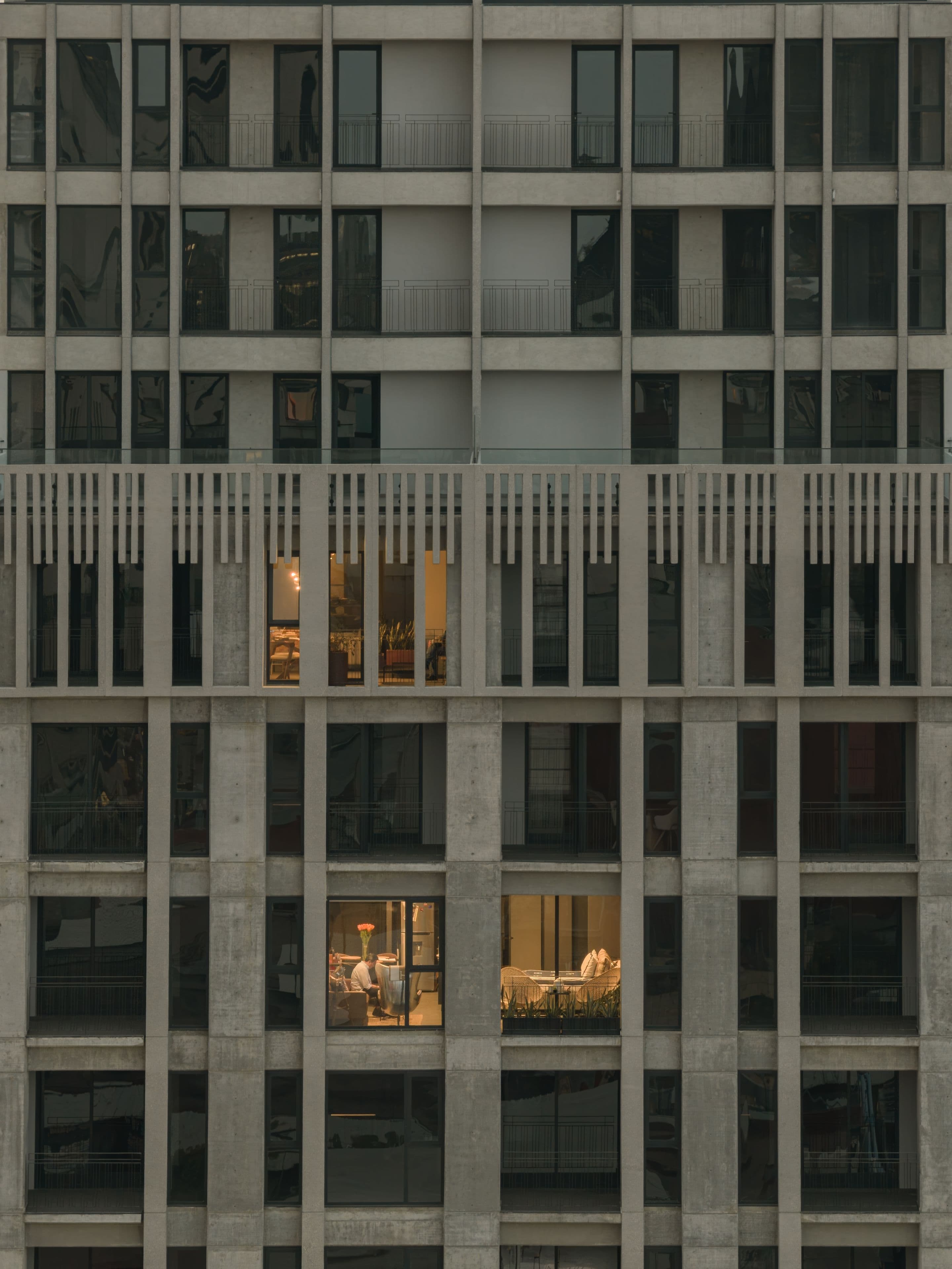 Un edificio de departamentos de concreto al anochecer, con tres cuartos encendidos: en uno, un hombre trabaja en su escritorio junto a unas flores.